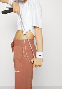 Hyppynaru, jossa on hopeiset kahvat ja mustat otteet, pidetään yhdessä kädessä. Valkoinen hikihattu Nike-logolla ranteessa. Rentoa valkoista t-paitaa ja ruskeita housuja.