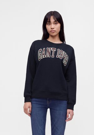 Femme aux cheveux bruns lisses portant un sweat-shirt noir avec "GANT 1949" en texte blanc et marron et un jean bleu, debout devant un fond blanc uni.