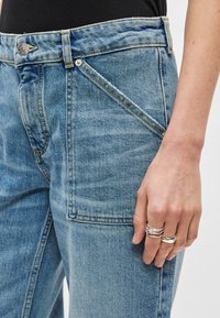 Jeans de ganga azul claro com cintura alta, apresentando um bolso frontal de remendo, costuras anguladas e detalhes em bolso com costura contrastante amarela.