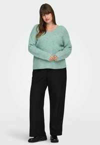 Šviesiai žalias, raštuotas megztinis su V formos iškirpte ir ilgomis rankovėmis, derinamas su juodomis plačiomis kelnėmis ir juodais batais.