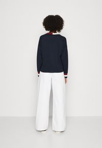 Granatowy sweter z okrągłym dekoltem, długimi rękawami i ściągaczami; zestawiony z białymi spodniami z szerokimi nogawkami. Minimalistyczny design bez widocznych wzorów.