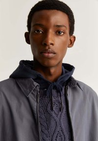 Veste navy avec fermeture éclair et col pointu, sur un pull en tricot texturé violet avec un motif en cablage, doté d'une capuche avec cordon de serrage.