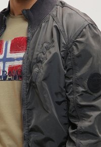 Grå bomberjakke med ribbet krave, sidelommer og logopatch på ærmet. Brugt over en lys beige t-shirt med grafisk print.