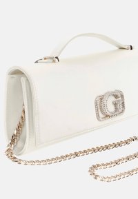 Bolso de mano de imitación cuero blanco con forma rectangular, correa de cadena dorada y un logotipo adornado con piedras brillantes en el frente.
