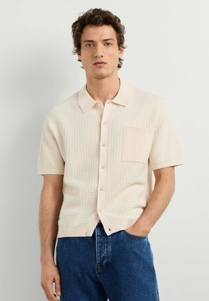Junger Mann mit kurzem lockigem Haar, der ein cremefarbenes kurzärmeliges Hemd mit Knöpfen und blaue Jeans trägt, steht mit einer Hand in der Tasche.