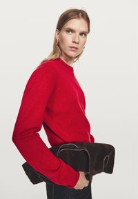 Červený pletený svetr s kulatým výstřihem a dlouhými rukávy; model drží tmavě hnědý semišový kabátek s hladkou texturou. Jednoduché pozadí.