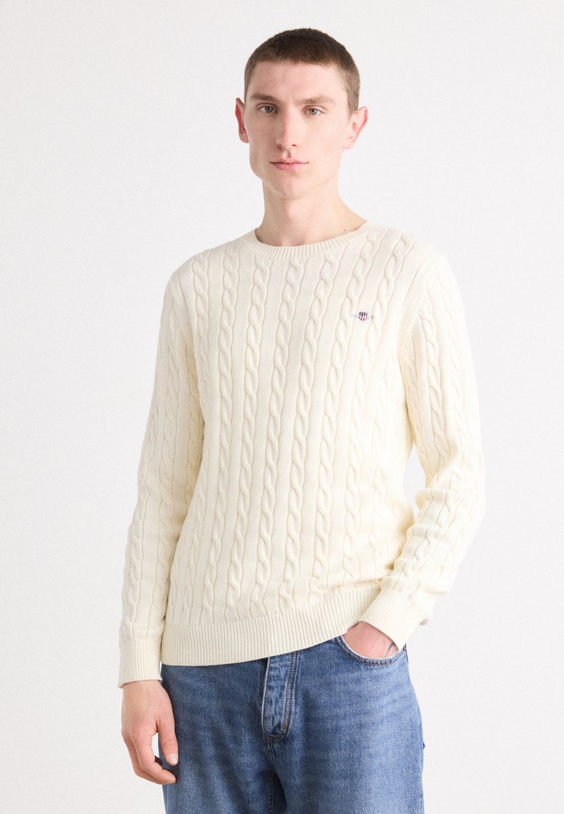 Cream cable-knit sweater with a round neckline, ribbed cuffs, and hem. Features a small logo on the left chest. Paired with blue jeans.