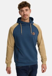 Sudadera azul con mangas raglán beige, cordón ajustable, bolsillos laterales y un detalle de logo circular en el pecho. Material suave de mezcla de algodón.