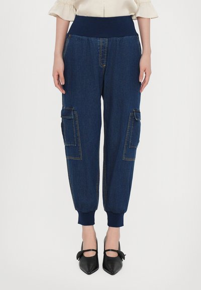 Pantalons jogger en denim bleu foncé avec des poches cargo aux genoux, des poignets élastiques et une large taille. Présente des surpiqûres dorées contrastantes.