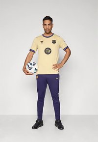Futbalový dres v žlto-námorníckej farbe s logom na hrudi, doplnený námorníckymi nohavicami. Model drží bielu futbalovú loptu s modrými detailmi.