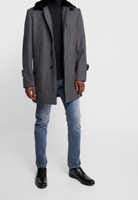 Grauer Mantel mit schwarzem Pelzkragen, Knopfverschluss, zwei Seitentaschen, kombiniert mit einem schwarzen Rollkragenpullover und hellblauen Jeans. Schwarze Schuhe.