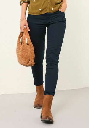Person geht und trägt dunkle enganliegende Hosen, braune Wildleder-Ankle-Boots und hält mit einer Hand eine tanfarbene gewebte Handtasche, während die andere Hand in der Tasche steckt.