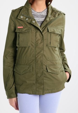 Mujer con chaqueta utilitaria verde oliva con cuatro bolsillos delanteros con solapa y pantalones lila claro, de pie con una mano en el bolsillo.