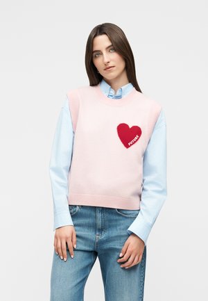 Mladá žena v modrej košeli a ružovom bezrukávniku s červeným srdcom a nápisom "BUDÚCNOSŤ", oblečená v modrých džínsach, stojaca s rukami pri bokoch.