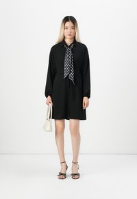Black dress with long sleeves and a waist tie, paired with a black and white polka dot scarf, white handbag, and black strappy sandals.