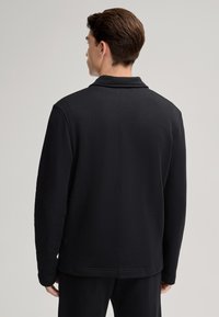 Indumento nero con colletto e maniche lunghe, realizzato in un materiale testurizzato, caratterizzato da una vestibilità rilassata e dettagli di cucitura minimi.