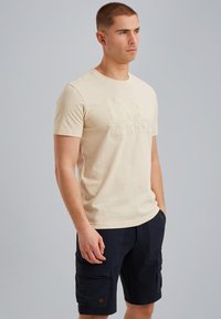 Beige puuvillane t-särk, mille ees on reljeefne logo, koos tumesiniste cargo-lühikestega, millel on suured küljetaskud ja kangast silt.