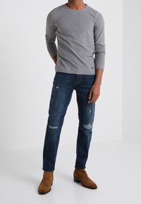 Graues Langarm-T-Shirt kombiniert mit dunkelblauen, ausgewaschenen Jeans und braunen Wildleder-Stiefeletten, vor einem schlichten weißen Hintergrund.