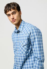 Camicia a quadri blu realizzata in tessuto leggero. Presenta due tasche sul petto con chiusura a bottone e un colletto a punta. Texture liscia.