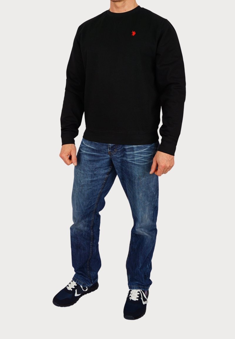 Homme portant une chemise noire à manches longues avec un logo rouge sur la poitrine, un jean bleu et des baskets noires avec des lacets blancs, debout devant un fond blanc.
