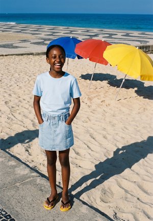 Glunderend meisje in een lichtblauw hemd en spijkerrok staat op een zandstrand nabij gele, rode en blauwe parasols met de oceaan op de achtergrond.