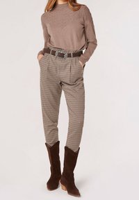 Brauner Strickpullover mit strukturiertem Muster, karierte beige und braune hochgeschnittene Hosen und braune Cowboystiefel aus Wildleder mit Absatz.
