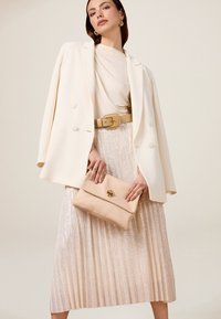 Blazer bianco sopra una maglia beige, cintura dorata e gonna plissettata metallica. Tiene una pochette beige con un pulsante dorato. Design semplice ed elegante.