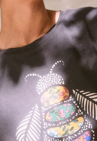 Gros plan d'une chemise sombre avec un motif abeille floral coloré, des ailes blanches et des antennes composées de petits cœurs.