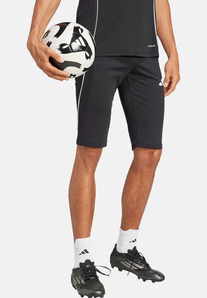 Schwarze Sportshorts mit weißen Seitenstreifen, kombiniert mit schwarzen Fußballschuhen und weißen Socken. Die Person hält einen schwarz-weißen Fußball.