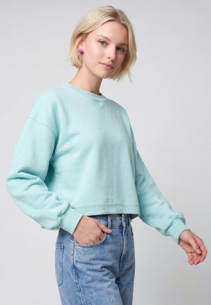 Jeune femme aux cheveux courts et blonds porte un sweat-shirt bleu clair, un jean bleu et des boucles d'oreilles rondes roses, debout devant un fond uni.