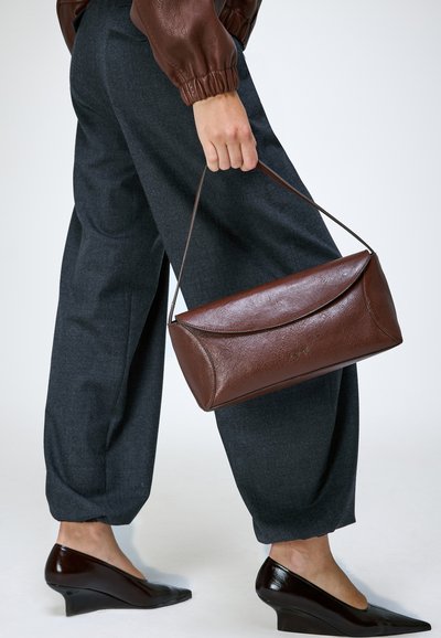 Personne portant un pantalon gris foncé et une veste en cuir marron tient un sac à main en cuir marron tout en portant des chaussures compensées marron.