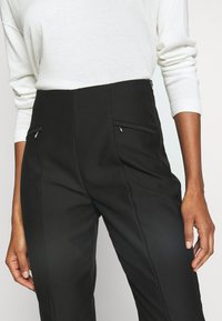 Personne portant un pantalon noir taille haute avec poches zippées à l'avant, associé à un haut blanc à manches longues avec manches retroussées.