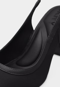 Μαύρο γυναικείο παπούτσι με λουράκι πίσω και ψηλό τακούνι, με επενδεδυμένη σεζούλα και στρογγυλεμένη μύτη, που δείχνει κοντινή λήψη της ραφής και υφής υλικού.