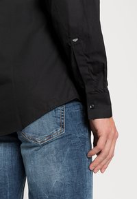 Camicia nera a maniche lunghe con una texture liscia, abbinata a jeans blu con un design sbiadito e cuciture a contrasto. Dettagli dei polsini abbottonati visibili.