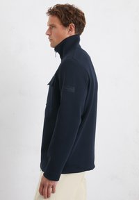 Donkerblauwe fleecejack met rits en hoge kraag, voorzien van een borstzak en een logo-opstuk op de mouw, met een soepele textuur en een ontspannen pasvorm.
