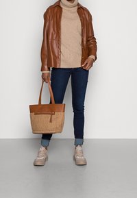 Vihreä nahkapaidan päällä on beige neulepaita, siniset farkut ja vaaleanbeige kengät. Hän pitää käsissään vaalean ruskeaa, kudottua olkalaukkua, jossa on vetoketju.