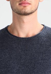 Dunkelgrauer, strukturierter Strickpullover mit Rundhalsausschnitt und sichtbaren Nähten. Nahaufnahme, die sich auf den Kragen und die Stoffdetails konzentriert.