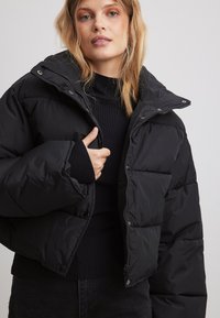 Melnā pūkaina jaka ar kvadrātu ielāpiem, ar rāvējslēdzēju un regulējamu kapuci. Mīksts audums ar ribotiem piedurknēm un īsu piegriezumu.