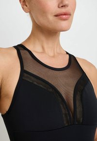 Maillot de bain noir avec des panneaux en maille au niveau du col, un design ajusté et une texture de tissu lisse. Les bretelles sont larges et soutenantes.