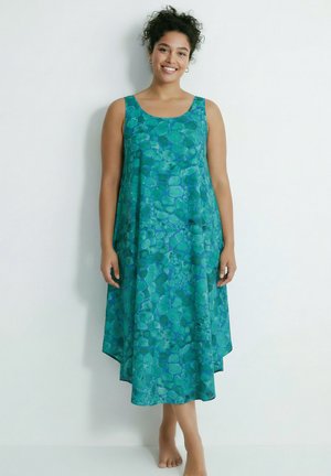 Smilende kvinde med krøllet hår iført ærmeløs, ankel-lang teal kjole med abstrakt cirkelmønster, stående barfodet op ad en hvid væg.