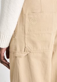 Beige cargo pants with a textured fabric. Features a side pocket with stitched detailing and a round logo embossed on the flap.