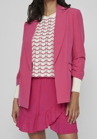 Rosa blazer över en vit tröja med vågmönster och en rosa volangkjol. Materialen verkar lätta, med en slät textur och skräddarsydd design.