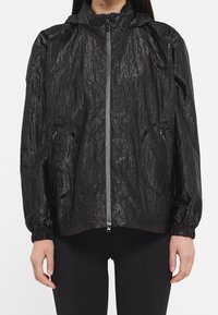 Kobieta w czarnej, pomarszczonej kurtce z zamkiem, z elastycznymi mankietami i zamykanymi kieszeniami, w połączeniu z czarnymi spodniami, stoi przodem.