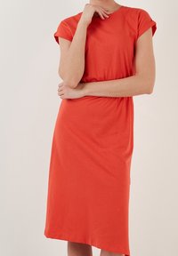 Mujer vestida con un vestido naranja brillante de longitud hasta la rodilla y mangas arremangadas, de pie con un brazo cruzado y la otra mano cerca de su mentón.