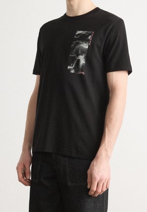 Czarny t-shirt z krótkim rękawem z białym i szarym abstrakcyjnym prostokątnym wzorem oraz małym czerwonym napisem na górnej lewej stronie klatki piersiowej.