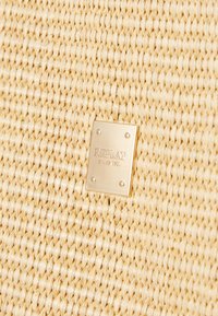 Woven straw material in a light beige color with a gold metal label featuring "REPLAY" and "SINCE 1981" attached. Textured surface pattern.