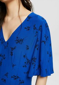 Robe sans manches bleue avec un décolleté en V profond, des manches flottantes et des broderies florales noires sur tout le tissu. Texture lisse.