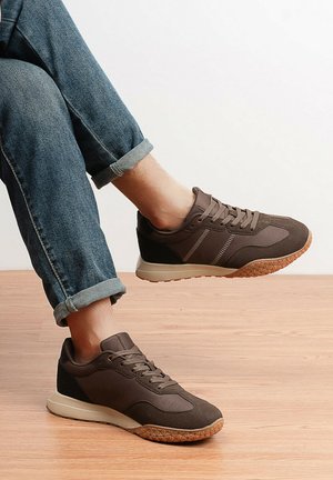 Personne portant un jean bleu retroussé et des baskets décontractées marron, debout sur un sol en bois devant un fond blanc uni.