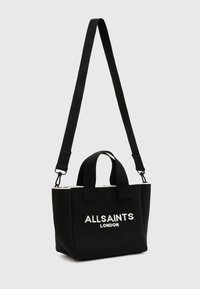 Borsa a tote nera in tela con due manici corti e una lunga tracolla regolabile. Presenta la scritta bianca "ALLSAINTS LONDON" e una cerniera bianca a contrasto.