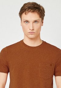 Bruine t-shirt met korte mouwen, gemaakt van gestructureerde stof, met een borstzak en een subtiel gestructureerd patroon. Ronde halslijn.